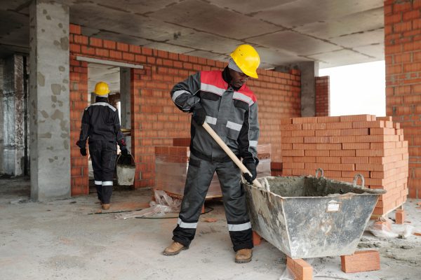 Mature builder mixing concrete in metallic basin against colleague carrying bucket with material for building brick wall